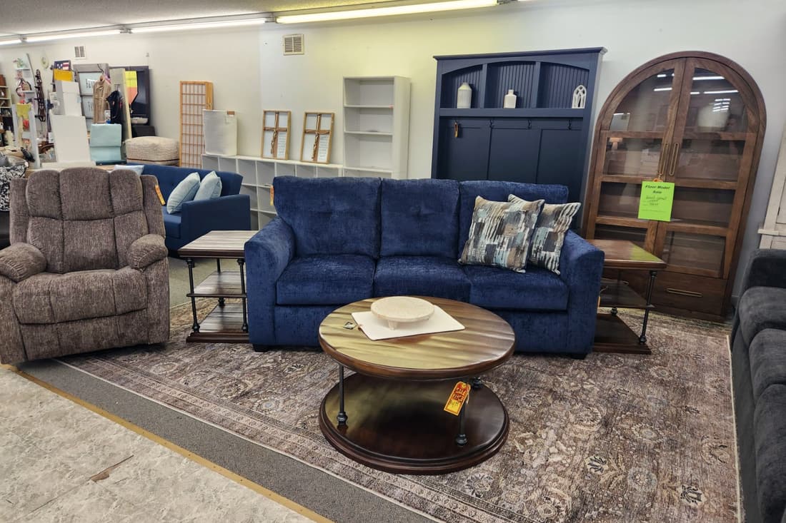 A navy sofa, a grey recliner and a round wood coffee table arranged on a patterned rug in the Thieves Market showroom
